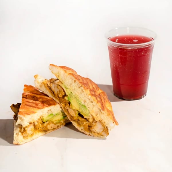 Grilled panini sandwich with avocado and chicken, alongside a red sparkling drink.